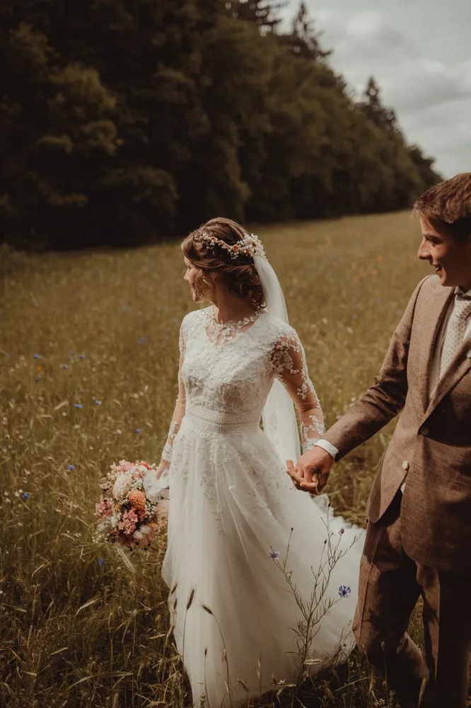 real-bride-review-geerdine-bruidshuis-diana – bruid Geerdine en haar bruidegom Wilbert staan samen in een zonnig landschap terwijl zij een elegante trouwjurk met kanten mouwen en lange sluier draagt en een kleurrijk bruidsboeket vasthoudt
