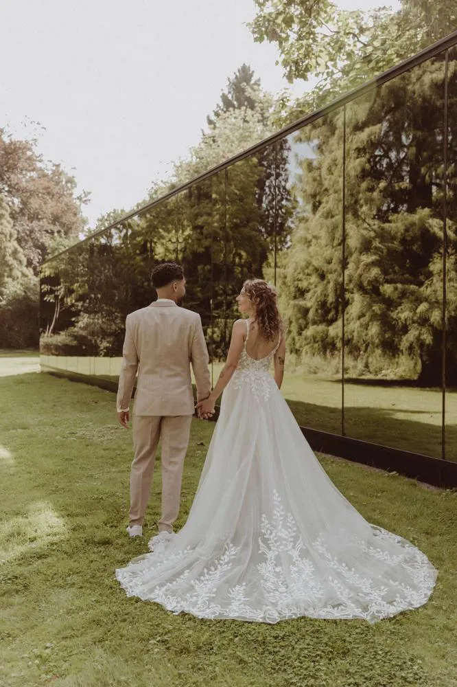 real bride stephanie van der linden – van der heijden – romantische A-lijn trouwjurk met lange sleep en bladapplicaties – bruid en bruidegom wandelen hand in hand langs een spiegelende muur in het groen