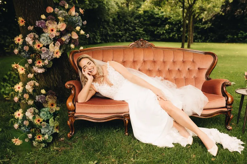 real-bride-review-dionne-van-schaick-foto-1-bruidshuis-diana – bruid lachend op vintage bank bij bloemen, tuinsetting.jpg