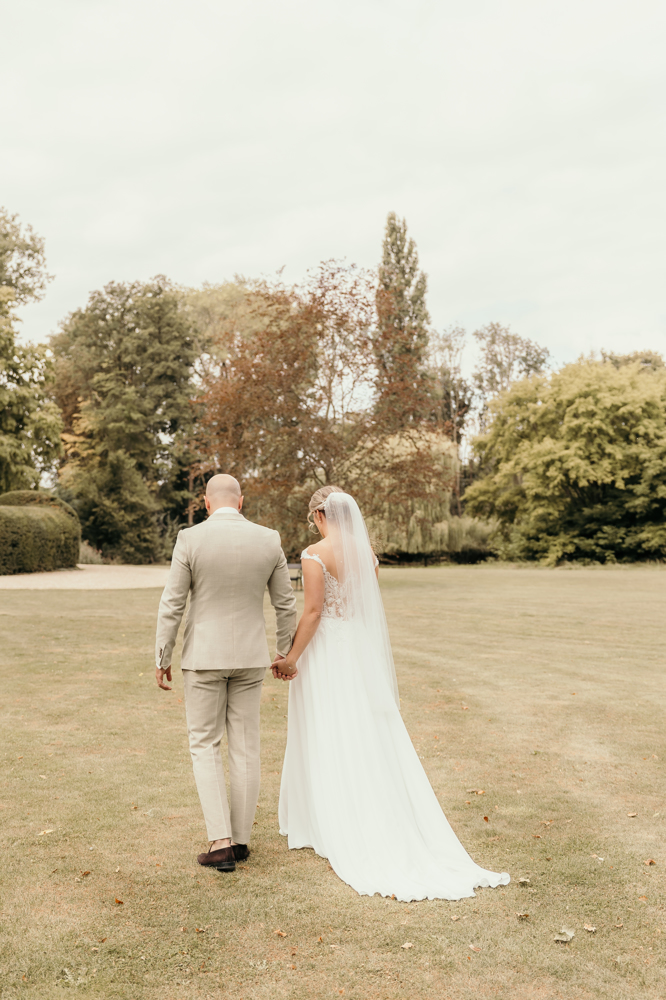 real-bride-review-babette-rosalle-bruidshuis-diana bruid Babette Rosalle hand in hand met haar partner op grasveld bij landhuis in trouwjurk met kanten lijfje
