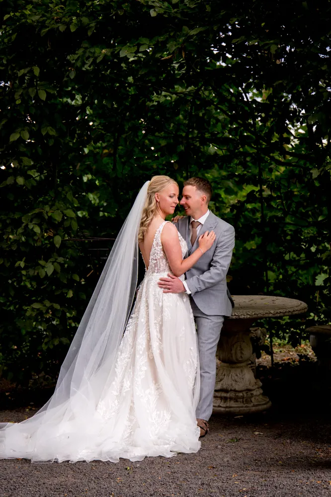 Real Bride Zohra & Jort – bruid en bruidegom omhelzen elkaar liefdevol in een groene tuinsetting; Zohra draagt een romantische A-lijn trouwjurk met verfijnde kanten details, open rug en lange sluier voor een elegante en tijdloze uitstraling.