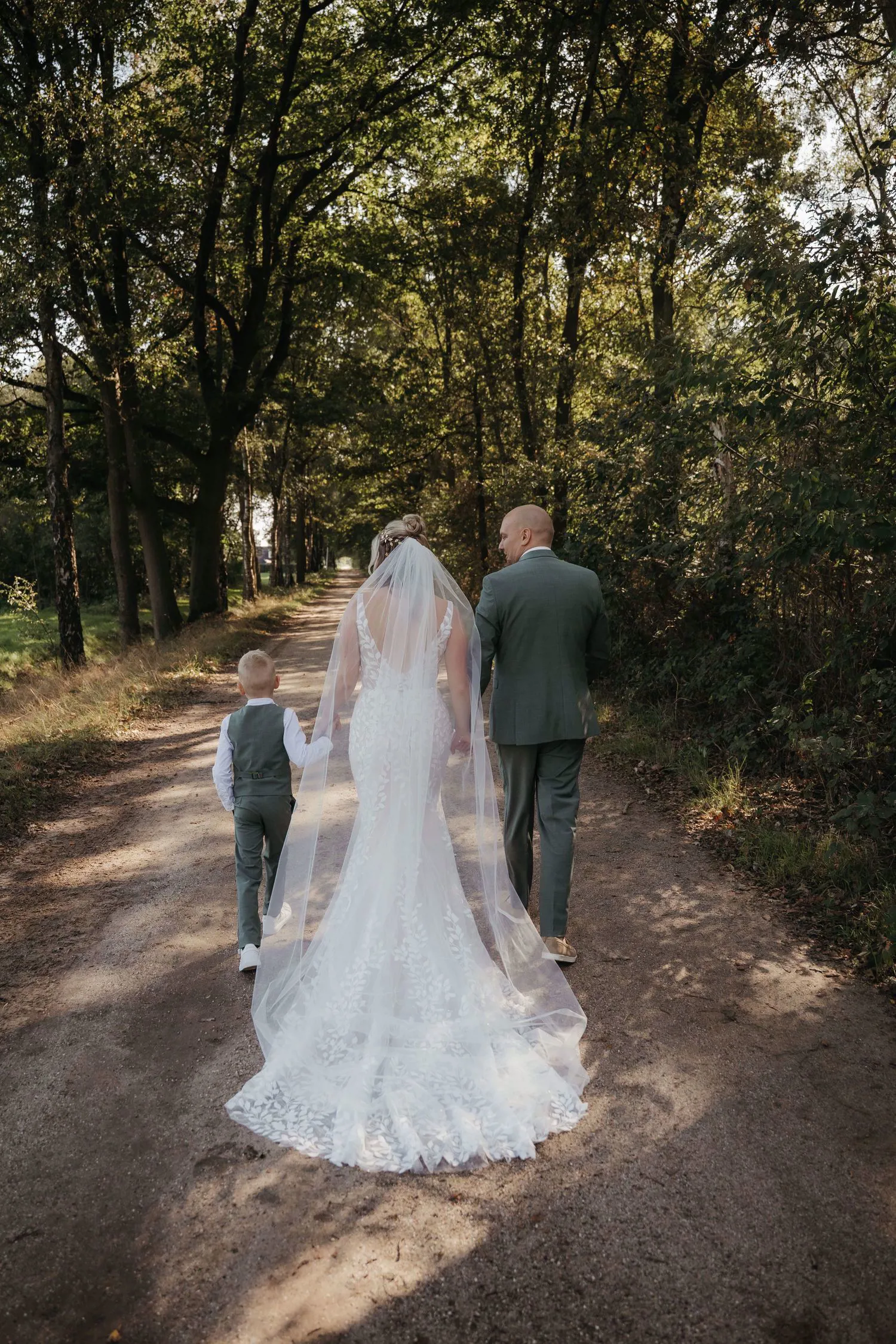 Real bride Lisa van Dooren loopt met haar gezin door een bos, in een fit & flare jurk met bladkant en sluier.